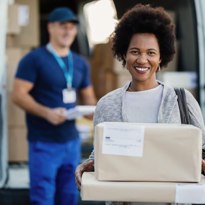 Portrait of happy black woman getting her packages delivered and looking at camera. A courier is standing in the background.